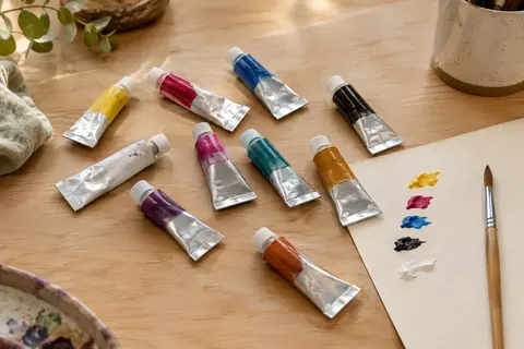 Dix tubes de gouache et un pinceau disposés sur une table d’atelier en bois clair, avec des touches de couleur.