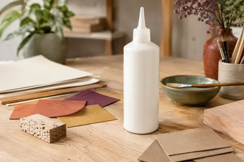 Flacon de colle vinylique blanche à embout fin posé près de papier, carton et bois sur une table claire.