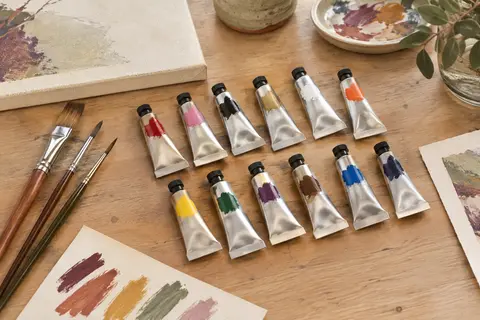 Douze tubes de peinture acrylique et trois pinceaux disposés sur une table d’atelier en bois clair.