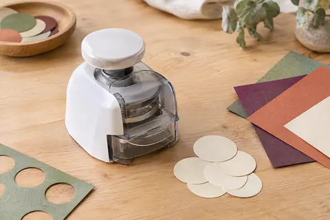 Perforatrice ronde blanche avec capot transparent, posée près de cercles découpés sur du papier crème.