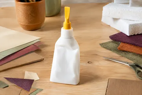 Flacon souple de colle vinylique avec spatule jaune, entouré de papier, carton et matériaux créatifs.